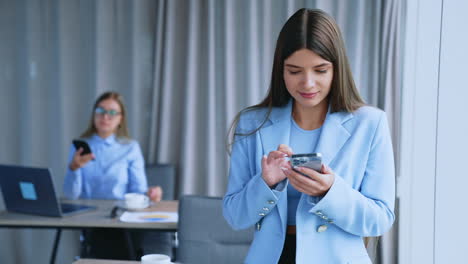 beautiful brunette lady with phone in her hands standing at the window. blonde colleague sitting at desk behind looking at her smartphone. blurred backdrop.