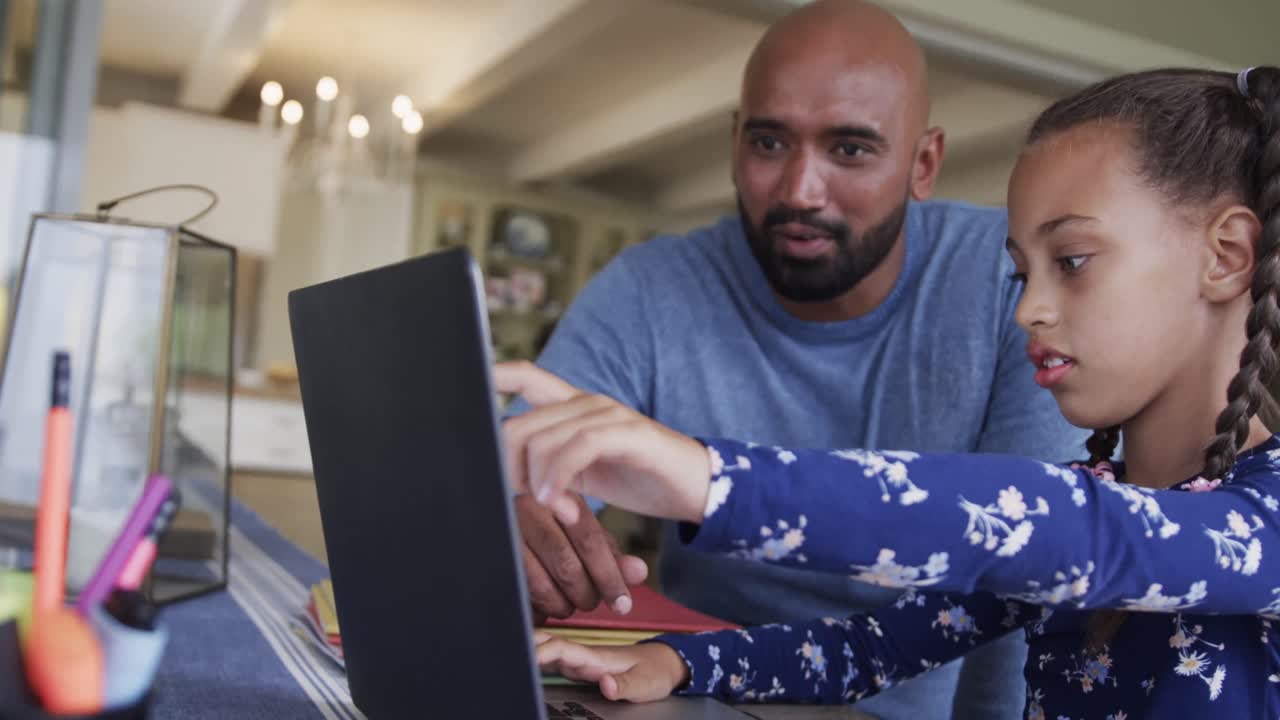 Happy biracial father and daughter learning online using laptop at table, slow motion