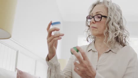 Senior-caucasian-woman-sitting-on-bed-wearing-glasses-looking-at-medicine-bottles,-slow-motion