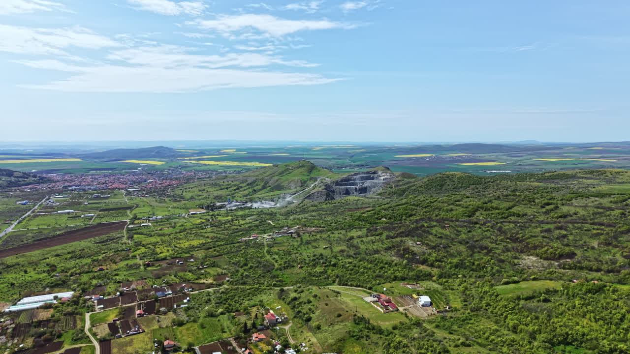 Vista de colinas verdes y un pueblo con una cantera de piedra al fondo, mostrando una mezcla de naturaleza e industria en el paisaje durante el día.