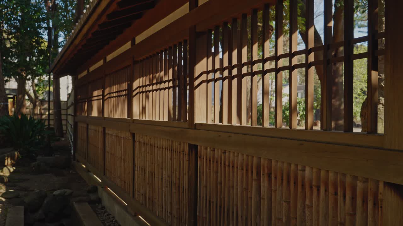 Una fluida panoramica verso destra che mostra una tradizionale recinzione in bambù e un traliccio in legno, catturando il gioco di luci e ombre in un'ambientazione di giardino tranquilla.