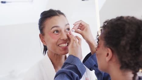 feliz pareja de lesbianas biraciales en albornoces aplicando máscaras debajo de los ojos en un baño soleado, cámara lenta