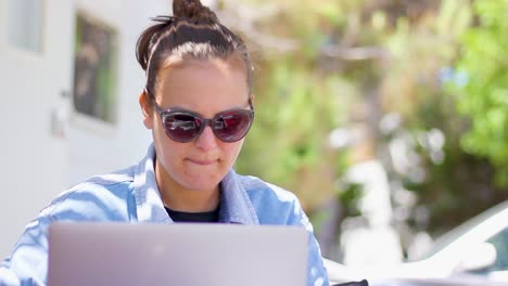 Young-woman-with-sunglasses-working-on-laptop-outside-caravan,-panning-shot