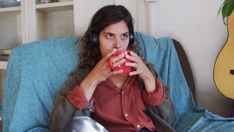 Relaxed-mixed-race-woman-wearing-headphones-sitting-drinking-tea-in-sunny-cottage-living-room