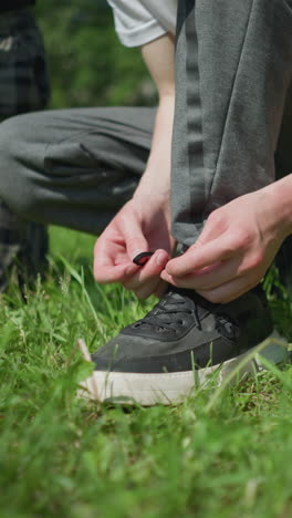 vista de la pierna de una persona ajustando el cordón de sus zapatos mientras se agacha en un campo de hierba, con una vista parcial de otra persona arrodillada en una pierna y una vista partial de alguien con ropa roja