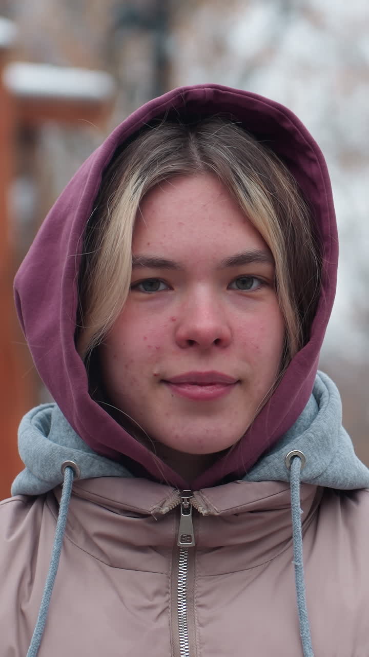 Giovane ragazza in abbigliamento invernale che tiene la catena dell'altalena, guarda in lontananza con un'espressione pensierosa, il viso ha lentiggini naturali, e uno sfondo sfocato di alberi e un ambiente esterno innevato.