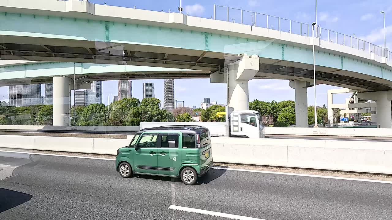 Vehicles traverse a sunlit Tokyo highway beneath an overpass. Trucks and cars move smoothly, highlighting urban transport dynamics