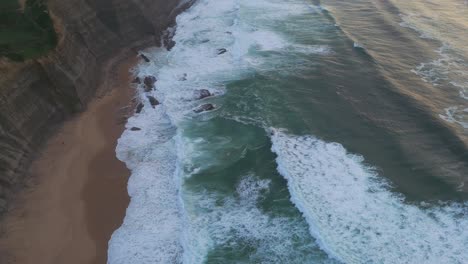 Vista-Aérea-Inclinada-Sobre-Las-Olas-En-La-Playa-De-Magoito,-Atardecer-Nublado-En-Portugal