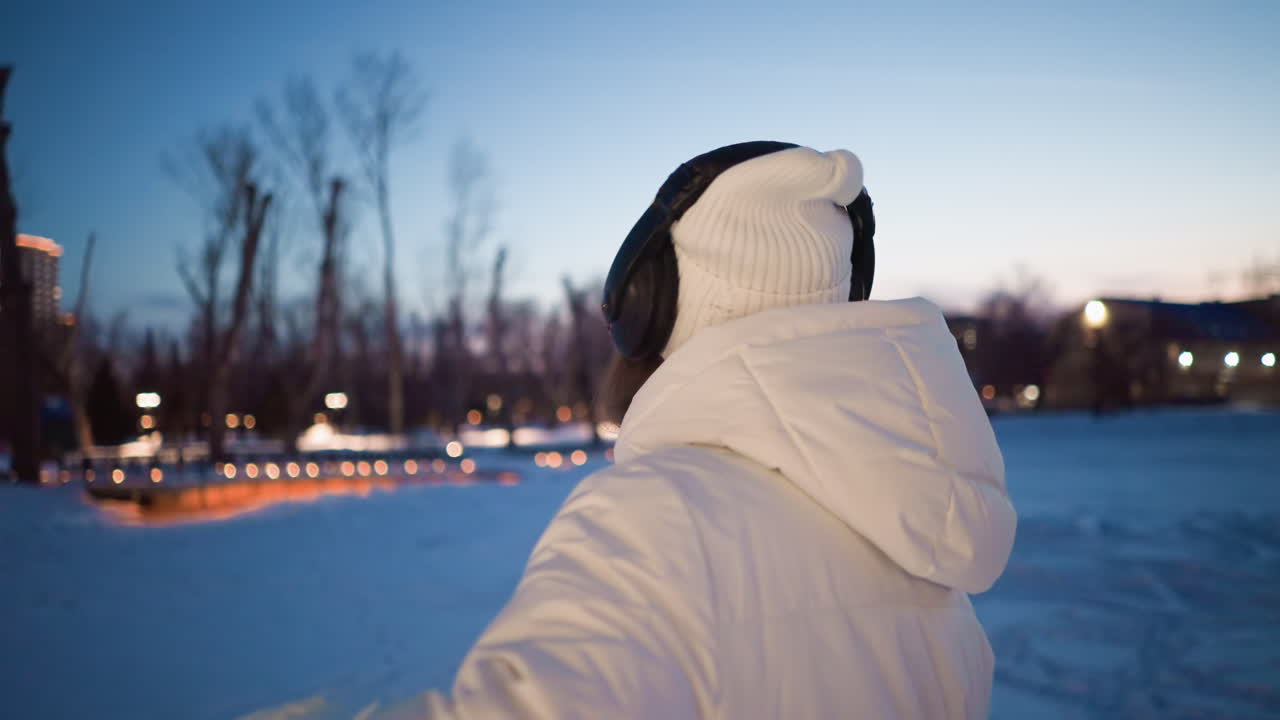 Student wearing white winter coat and headphones moves body in rhythmic dance outdoors on snowy ground at twilight with blurred trees and festive lights in background conveying joyful motion