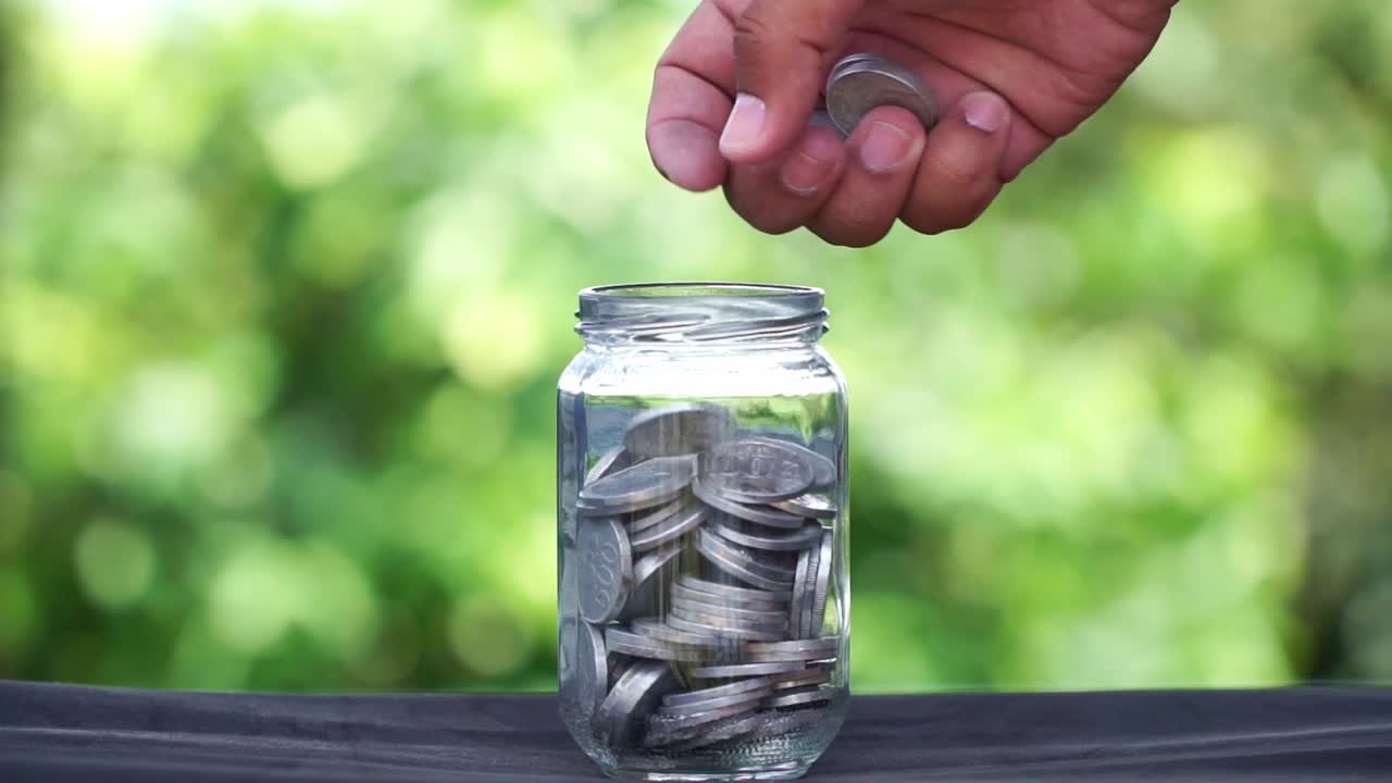 A human hand is inserting coins into a glass container that is half filled with coins.