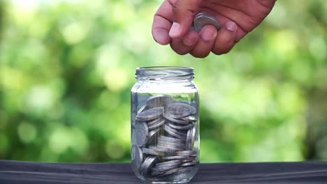 a human hand is inserting coins into a glass container that is half filled with coins.