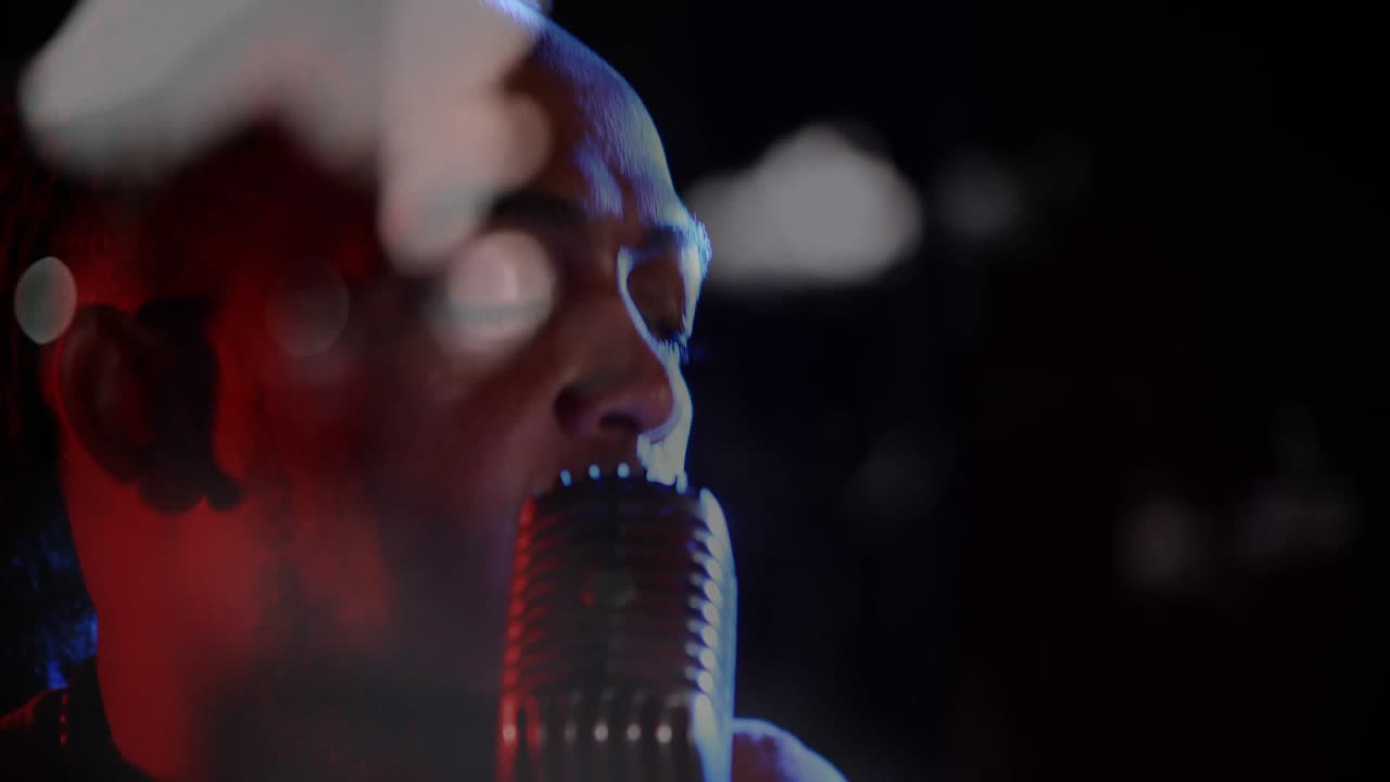 Camera pulling back, male singer leaning into studio mic, revealing face and dog-tag while singing