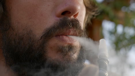 primer plano de un joven barbudo inhalando y exhalando un cigarrillo electrónico