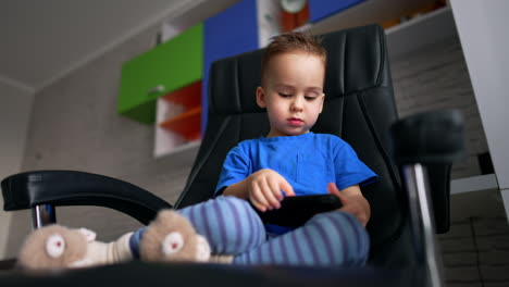 lovely caucasian toddler boy sits in a big chair. cute baby is focused on the phone in his hands. low angle view.