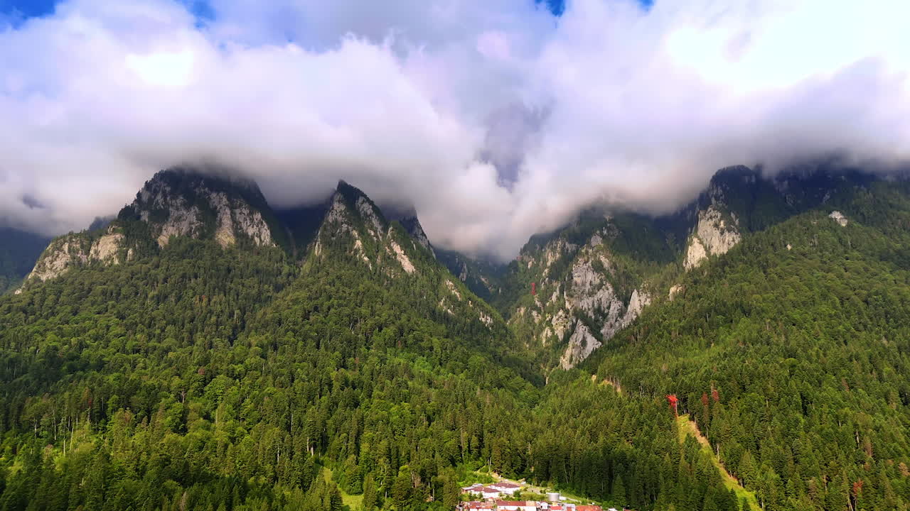 Alberi altissimi sovrastano le montagne, con nuvole basse all'orizzonte. Un piccolo villaggio è immerso nella valle sottostante, circondato da rigogliose foreste verdi in pieno giorno.