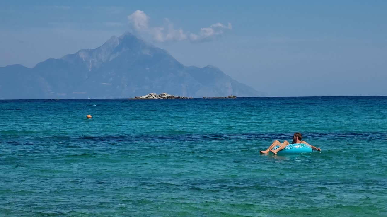 A man drifts on a blue inflatable ring in the turquoise waters of Kriaritsi Beach, Greece, soaking up the sun and carefree summer vibes on a bright, cloudless day
