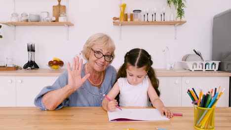 abuela y nieta mirando la cámara durante una videollamada mientras dibujan juntas sentadas en la mesa de la cocina