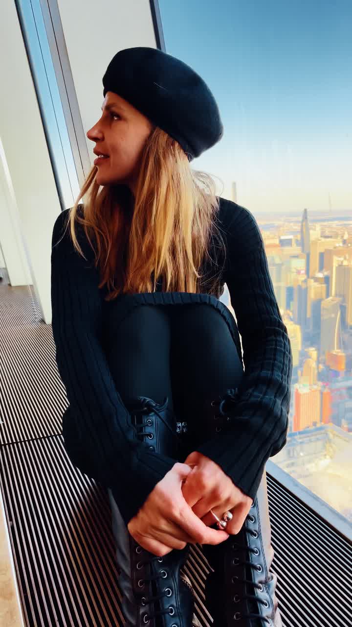 Porträt einer Frau am Fenster mit Blick auf die Skyline von Manhattan. Sitzende Frau mit schwarzem Barett und Kleid blickt in die Kamera nahe einem Fenster mit der Skyline von Manhattan im Hintergrund.