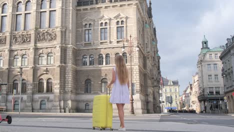 a young beautiful girl with a yellow suitcase is walking on a city street from behind. back view of a girl traveler walking and pulling a suitcase