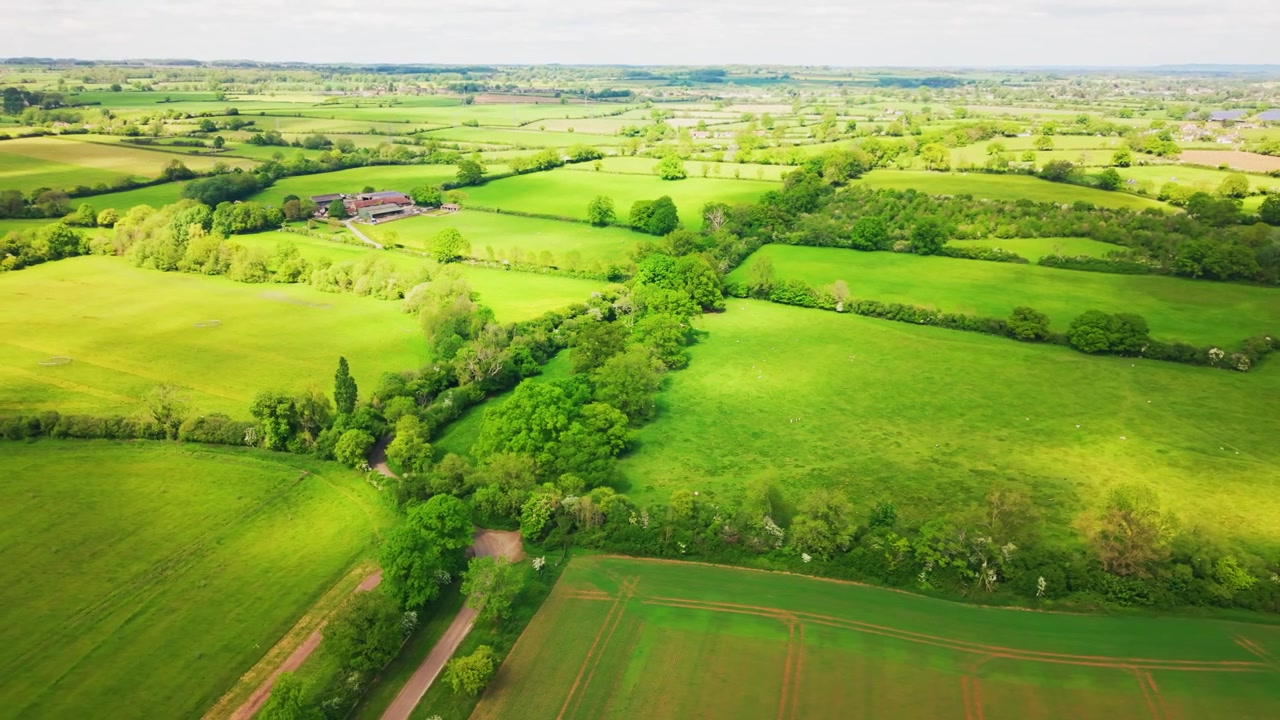 Wide aerial drone footage over green farmland, pasture fields, hedgerows, woodland, farm buildings, country lanes, and the surrounding English countryside.