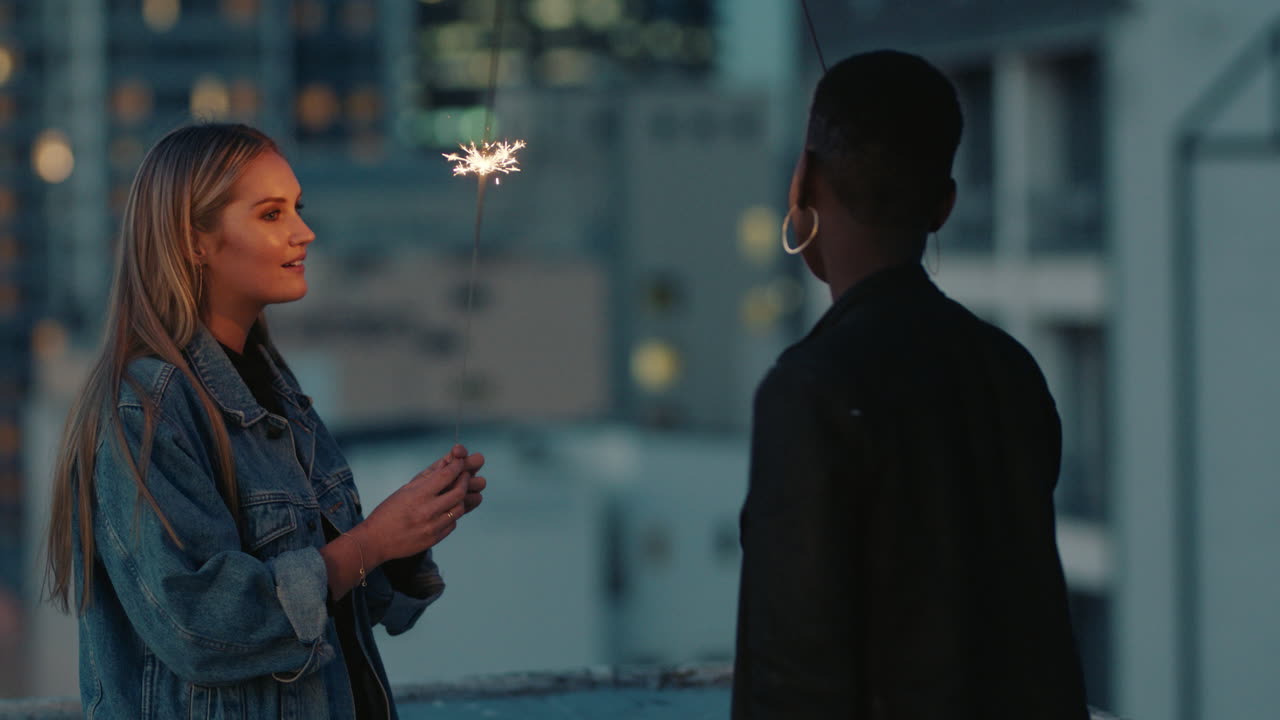 아름다운 젊은 여성 친구들은 일몰 때 옥상에서 새해 전야를 축하하는 폭죽을 들고 휴일 파티 축하를 즐기고 있습니다.