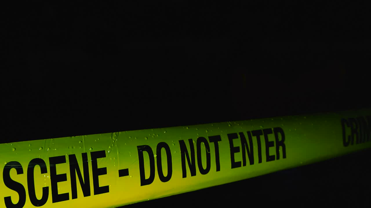 Yellow colored police line Crime Scene tape in front of a dark night rainy background while raindrops fall on the in ribbon in slow motion, footage pans over the forensics investigation tape