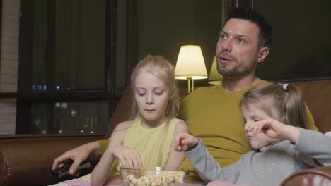 Papa-Mit-TV-Fernbedienung-Und-Seine-Beiden-Kleinen-Töchter-Essen-Popcorn-Und-Schauen-Sich-Einen-Film-An,-Während-Sie-Nachts-Zu-Hause-Auf-Dem-Sofa-Sitzen-1