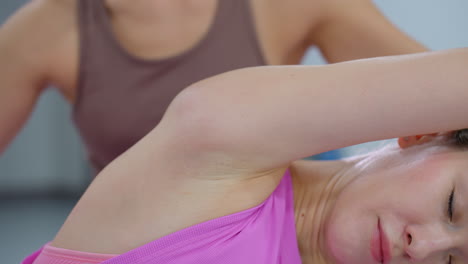 close-up of woman in pink performing lounge stretch with eyes closed, focusing on flexibility and relaxation, trainer in brown outfit stands nearby, guiding the workout session