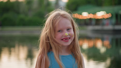 portrait view of beautiful young girl with flowing hair smiling with bright green paint dots on nose against softly blurred riverside park lights backdrop during warm golden evening art session