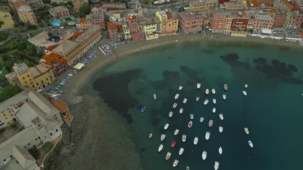 Sestri Levante, a picturesque town in Liguria, Italy, is captured from above, showcasing colorful waterfront buildings, a serene harbor with boats, and clear blue waters along the sandy beach