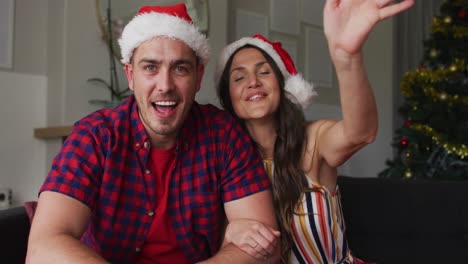 una feliz pareja caucásica con sombreros de santa sentada en el sofá, haciendo gestos, haciendo una videollamada