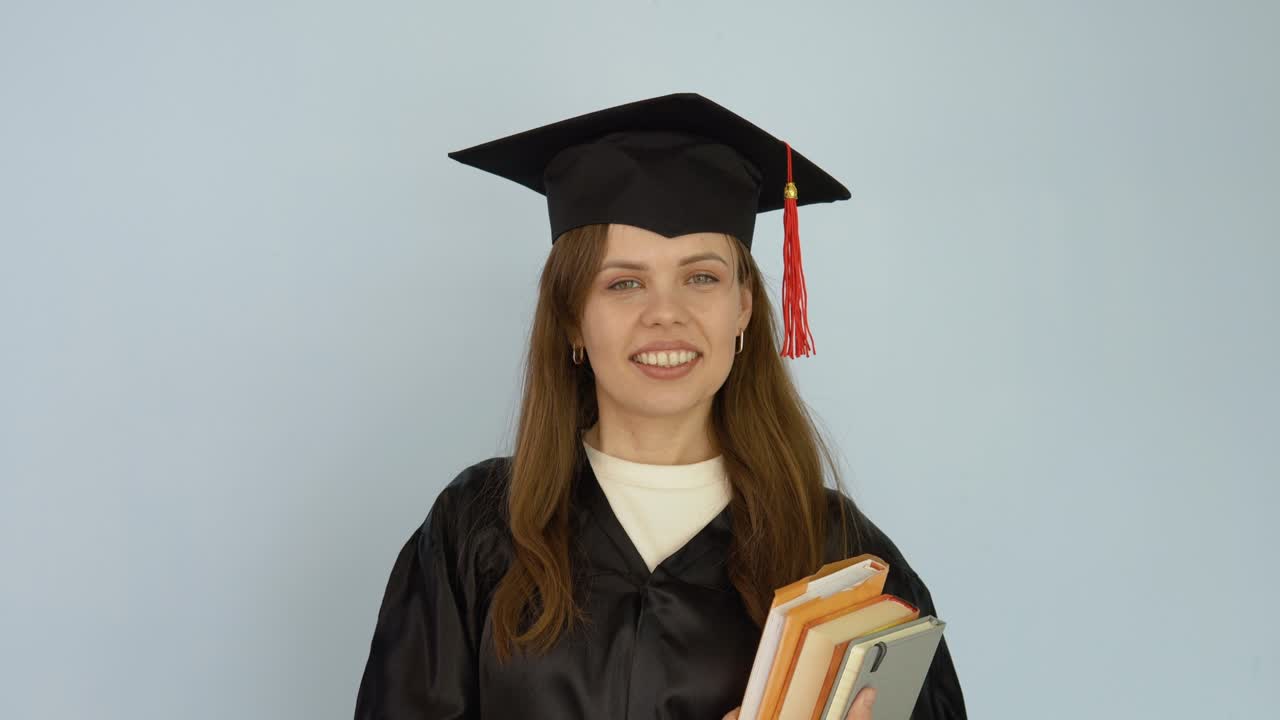 una joven mujer caucásica con una túnica negra y un sombrero de maestro se mantiene erguida sosteniendo libros de texto y mostrando un buen comportamiento con el dedo gordo hacia arriba. el estudiante mira a la cámara sobre un fondo blanco