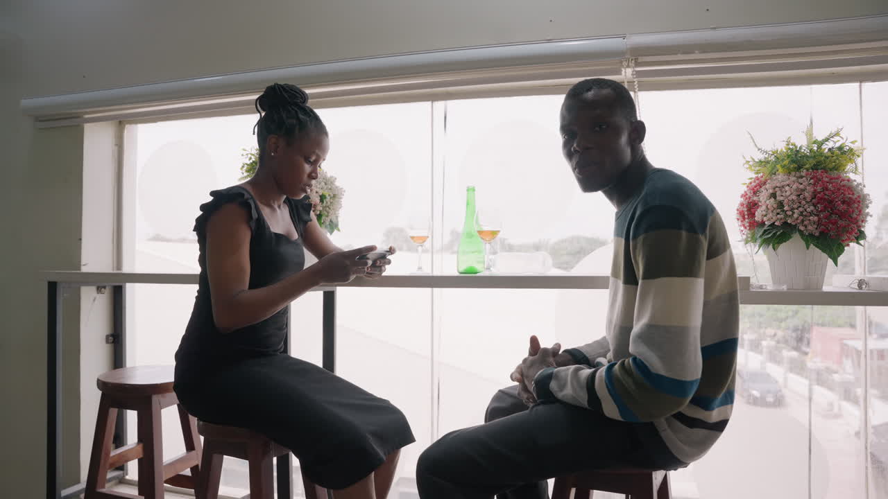 Black couple on cozy window date sharing quiet conversation over coffee by bright city window with bouquet on sill woman checking phone while man watches, gentle gestures and tender gazes, intimate