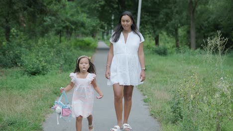 a pregnant woman and her young daughter, both dressed in white, walk hand-in-hand through a grassy park. the scene captures a serene moment of family bonding in nature, surrounded by trees.