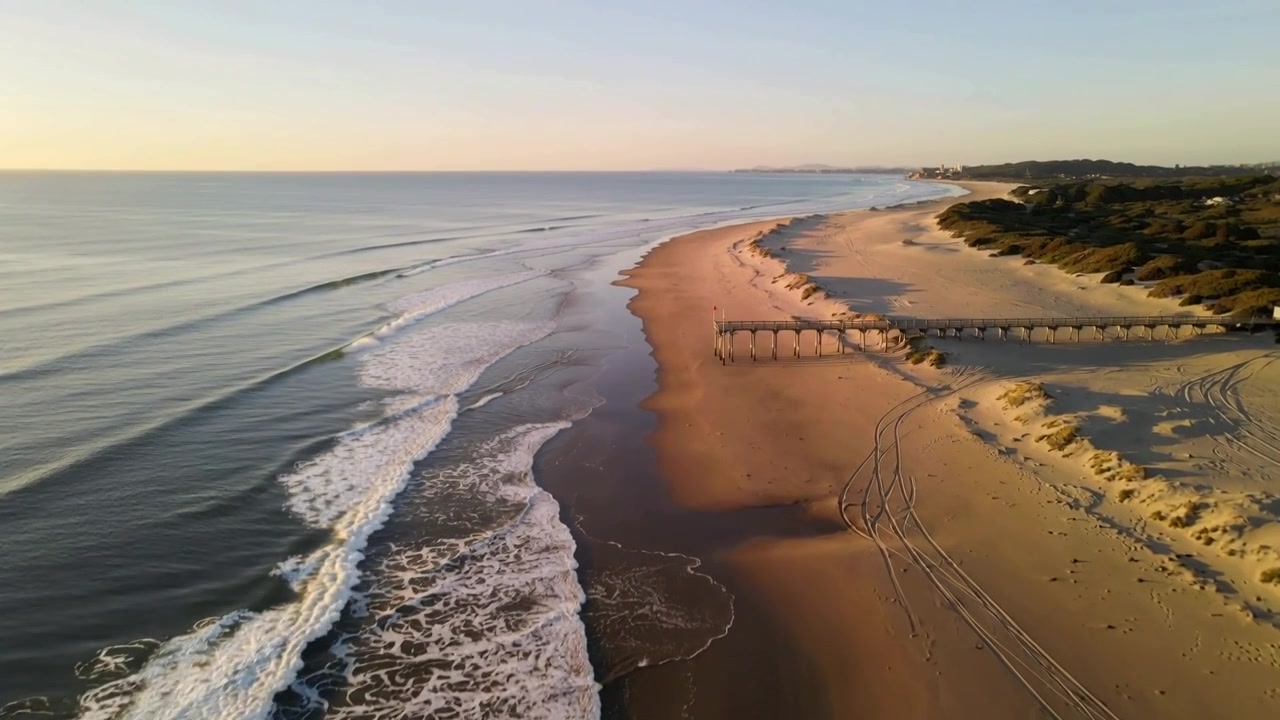 Zwevende drone onthult lange zandkustlijn bij laagstaande zon, met houten pier en bandensporen