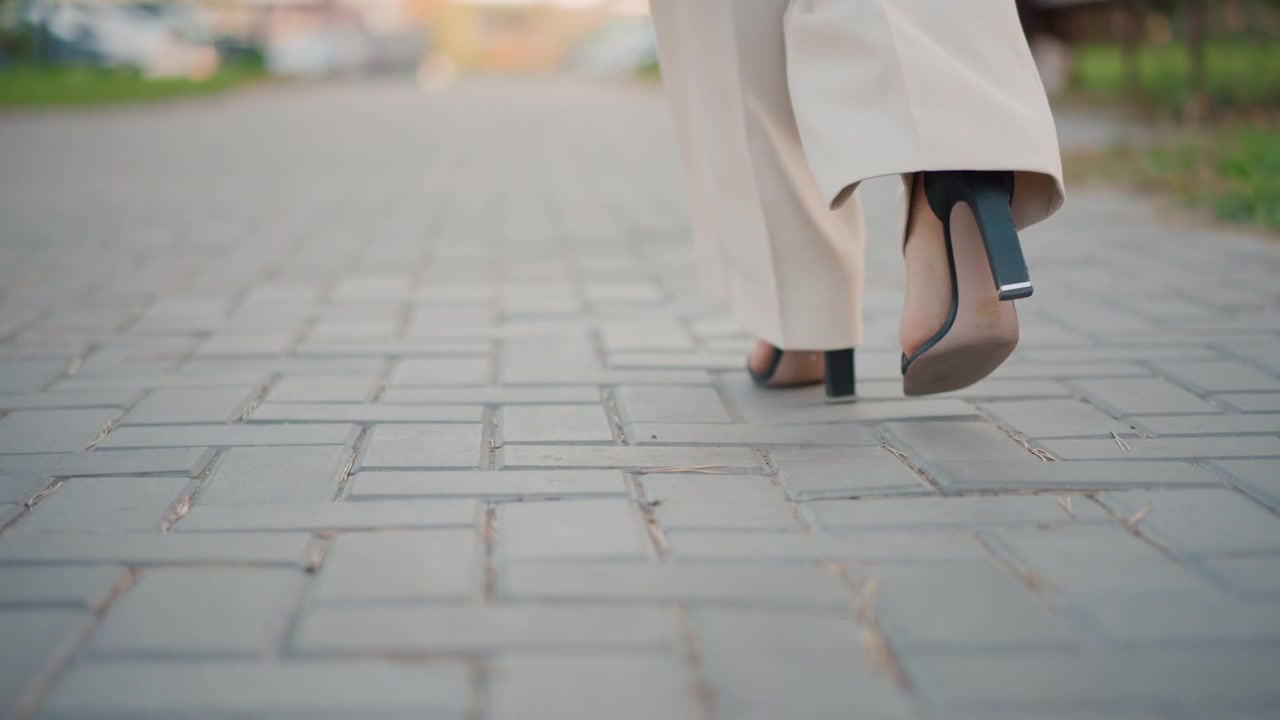 Rear low-angle view shows lower legs and high heels in mid-stride on paved sidewalk, with light skirt hem swaying, heel lift, and steady forward movement in soft daylight, with heel, walking