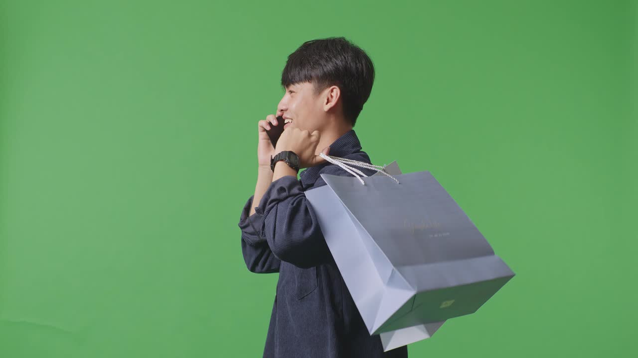 초록색 화면 앞에서 는 동안 전화에 응답하고 휴대전화로 대화하는 쇼핑 가방을 가진 아시아 쇼핑맨의 측면 시각