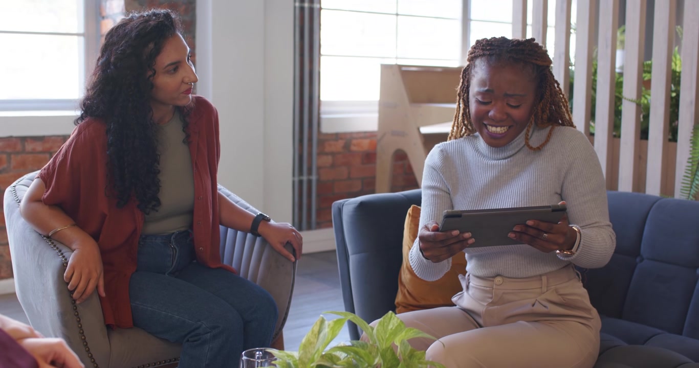 Female coworkers meeting in loft maroon blazer initiating, tablet and laptop showing content