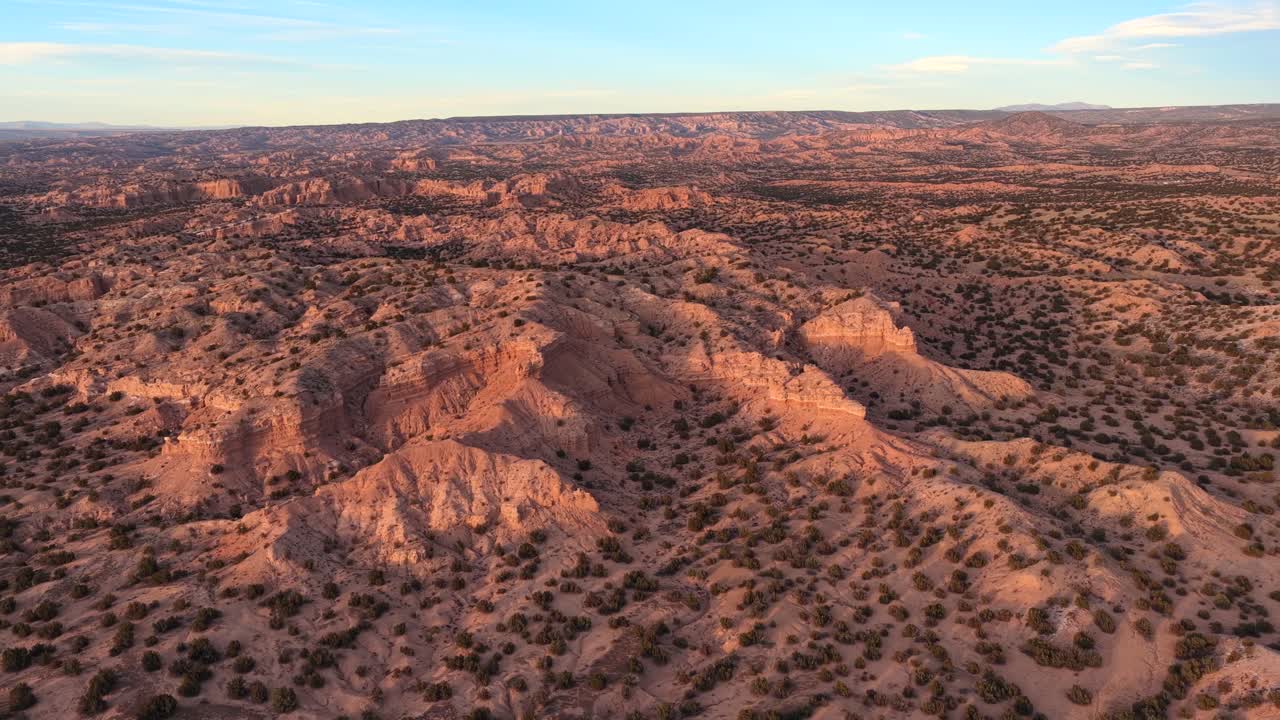A wide aerial perspective reveals the sharp textures and deep gullies of eroded sandstone badlands. The ancient high desert terrain features layered rock and sparse green shrubs in northern New Mexico