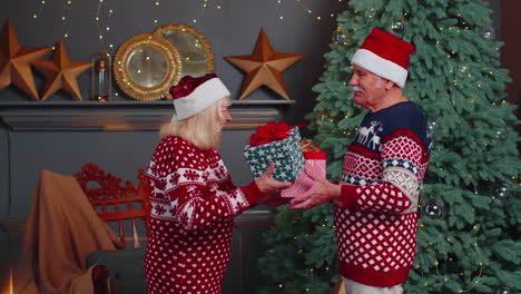 romantic senior couple presenting gift boxes to each other at home near decorating christmas tree