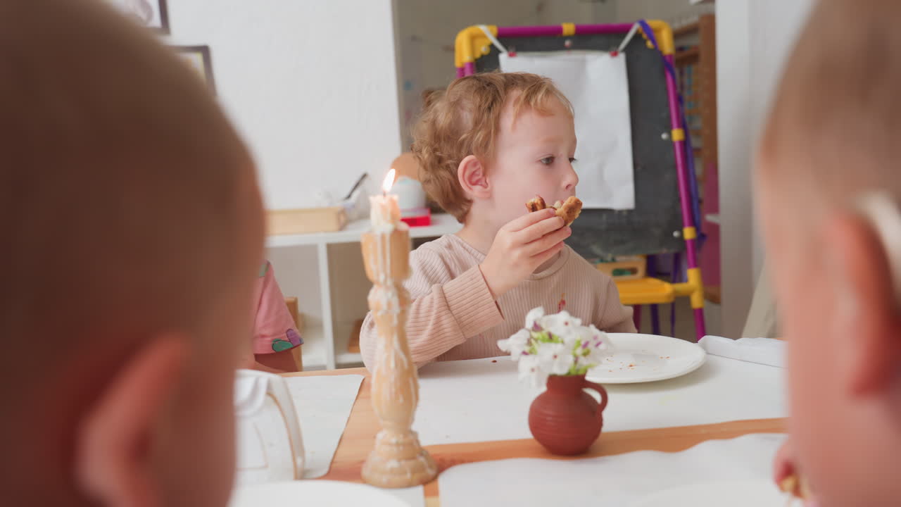 미취학 아동 간식 시간에 아이들이 음식을 먹는 모습 클로즈업, 보청기를 낀 남자아이가 음식을 받아 촛불 근처에서 빵을 씹고, 또래 아이들이 가장자리에서 지켜보는 따뜻하고 포용적인 교실 풍경