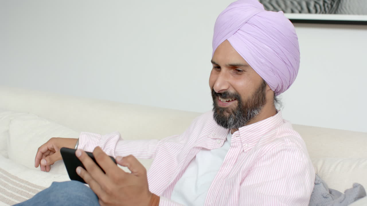 Uomo sorridente con turbante che usa il tablet mentre si rilassa sul divano a casa.