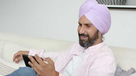 smiling man with turban using tablet while relaxing on couch at home