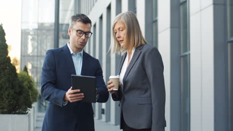 businessman showing project and new ideas on tablet device to woman boss who holds coffee to go in hands while they stand in the street