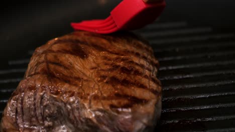 Extreme-close-up-shot-of-a-delicious-fried-steak-in-the-hot-pan-being-brushed-with-oil-with-a-red-brush-for-a-delicious-dinner