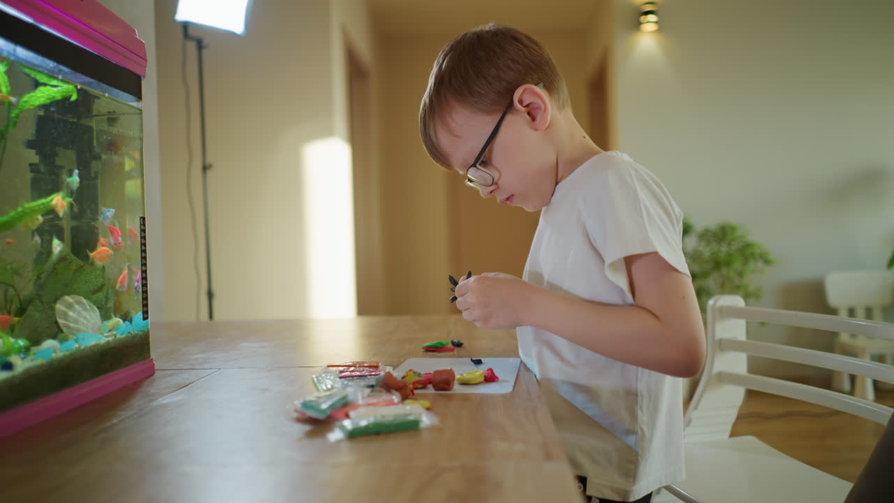 Boy in glasses sitting at table creating colorful clay figures with concentration, surrounded by scattered modeling clay pieces, aquarium with tropical fish in background, home interior, sunlight atmosphere