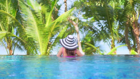 Vista-Trasera-De-Una-Mujer-Con-Sombrero-Grande-Y-Traje-De-Baño-Rosa-En-El-Borde-De-La-Piscina