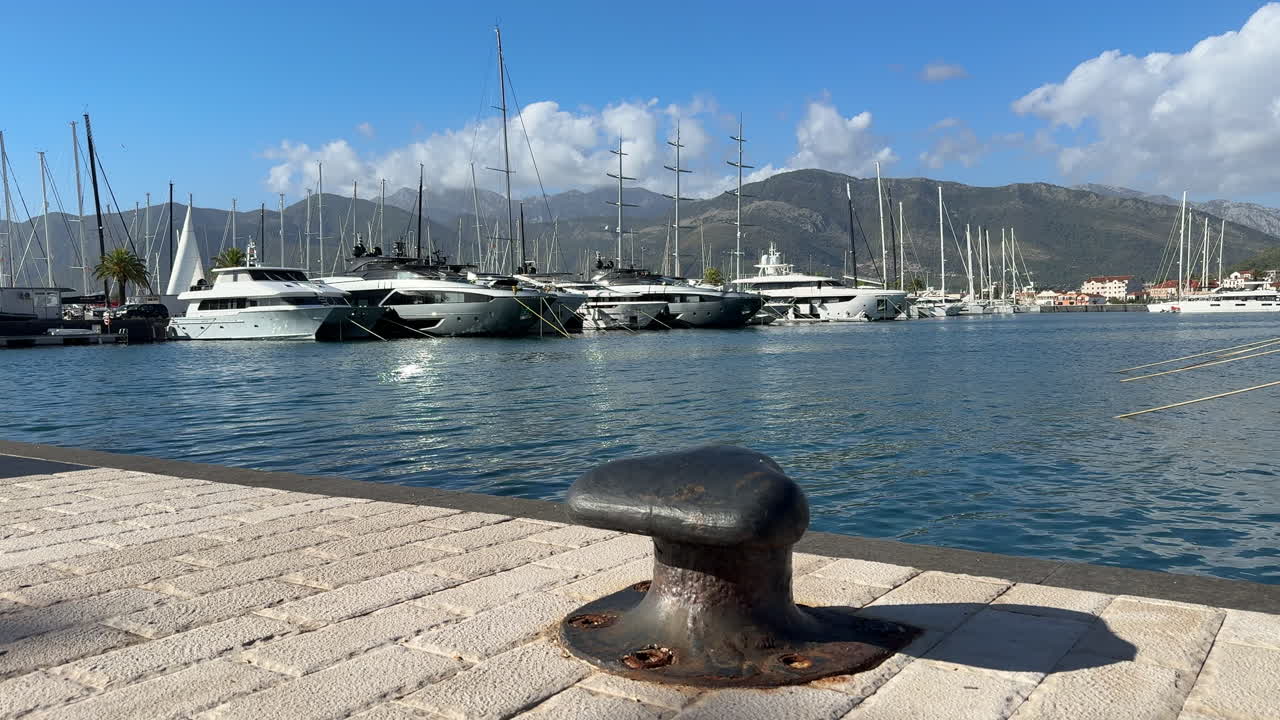 Grosso primo piano di una bitta da ormeggio con yacht di lusso e barche a vela a Porto Montenegro, incorniciata da acqua blu e montagne costiere. Per temi come viaggi di lusso, stile di vita nautico, turismo e scene di porticcioli turistici