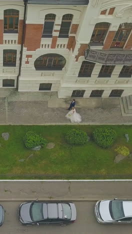 groom and bride walk along empty street past elegant restored building with lawn on autumn day upper view
