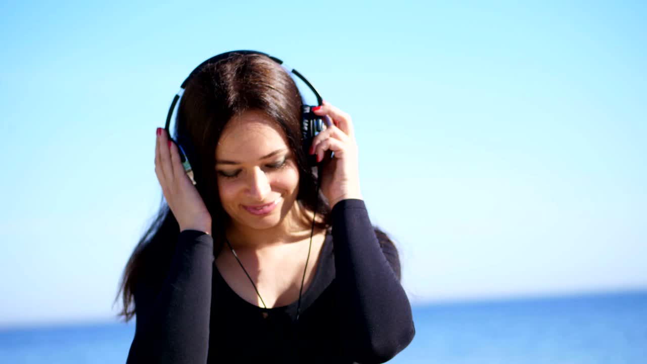 여름, 바다의 배경에. 긴 어두운 머리카락을 가진 아름다운 여성, 큰 헤드폰을 입고, 야외에서 음악을 듣고, 춤을 추고. 헤드폰으로 음악을 듣는 여성